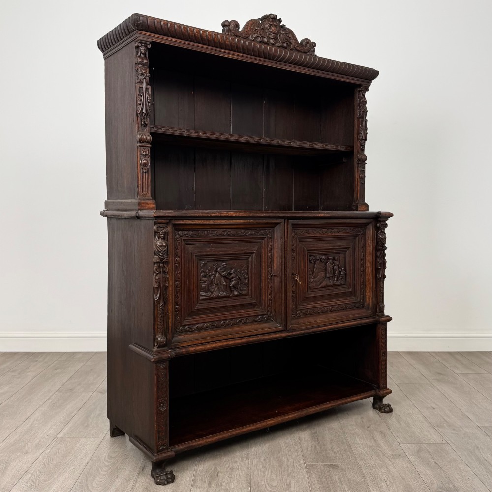 A 19th-century dark oak carved cupboard featuring an open top shelf, carved lion cresting, relief-carved panel doors with figures, and an open lower base. View IX