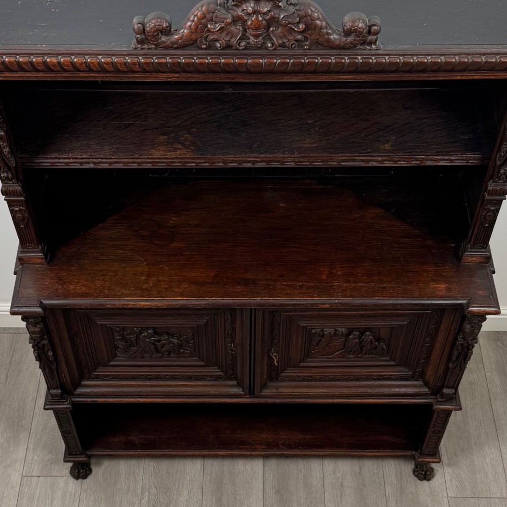 A 19th-century dark oak carved cupboard featuring an open top shelf, carved lion cresting, relief-carved panel doors with figures, and an open lower base. View X