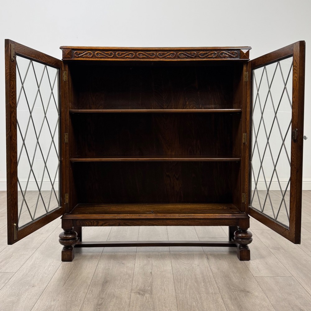 An Edwardian dark oak display cabinet with double diamond-leaded glass doors, a carved scroll frieze along the top, interior wooden shelves, and bulbous turned legs connected by a stretcher. View II