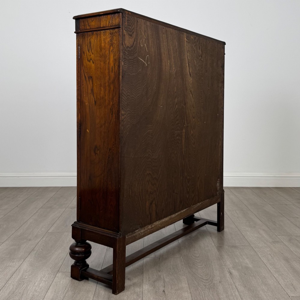 An Edwardian dark oak display cabinet with double diamond-leaded glass doors, a carved scroll frieze along the top, interior wooden shelves, and bulbous turned legs connected by a stretcher. View V
