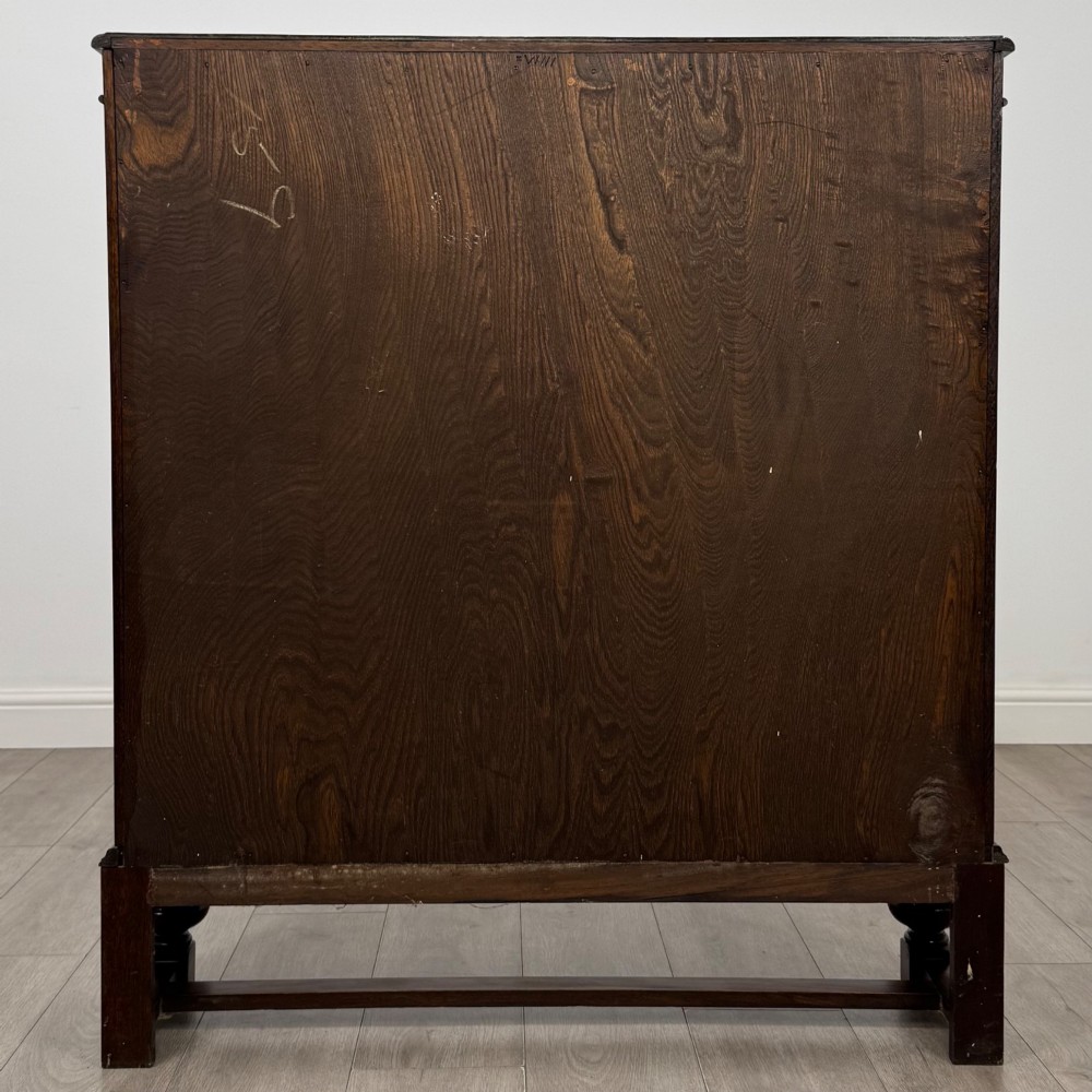 An Edwardian dark oak display cabinet with double diamond-leaded glass doors, a carved scroll frieze along the top, interior wooden shelves, and bulbous turned legs connected by a stretcher. View VI