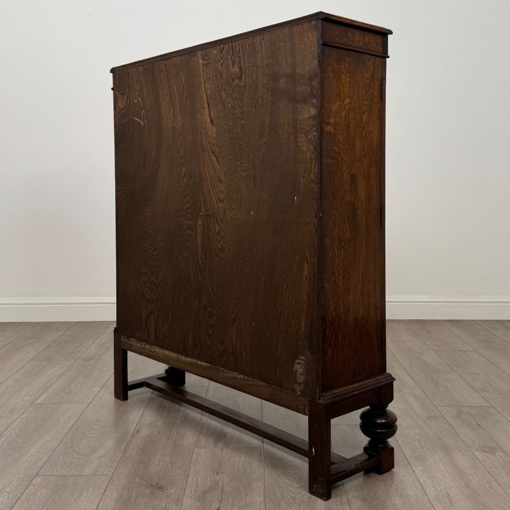 An Edwardian dark oak display cabinet with double diamond-leaded glass doors, a carved scroll frieze along the top, interior wooden shelves, and bulbous turned legs connected by a stretcher. View VII