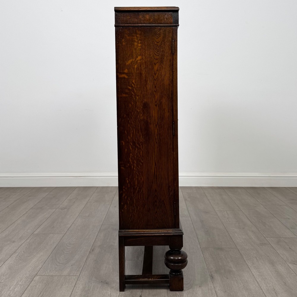 An Edwardian dark oak display cabinet with double diamond-leaded glass doors, a carved scroll frieze along the top, interior wooden shelves, and bulbous turned legs connected by a stretcher. View VIII