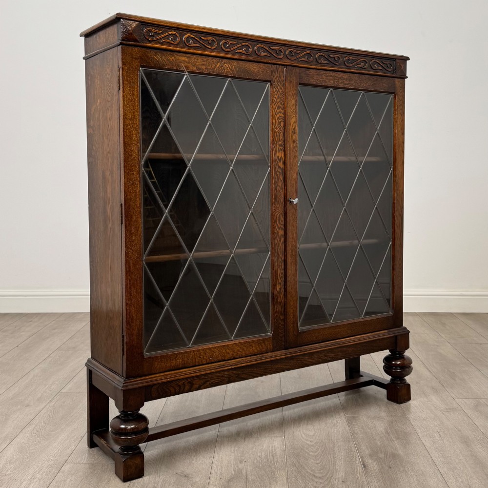 An Edwardian dark oak display cabinet with double diamond-leaded glass doors, a carved scroll frieze along the top, interior wooden shelves, and bulbous turned legs connected by a stretcher. View IX