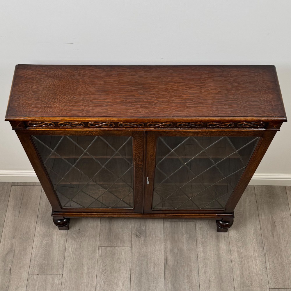 An Edwardian dark oak display cabinet with double diamond-leaded glass doors, a carved scroll frieze along the top, interior wooden shelves, and bulbous turned legs connected by a stretcher. View X