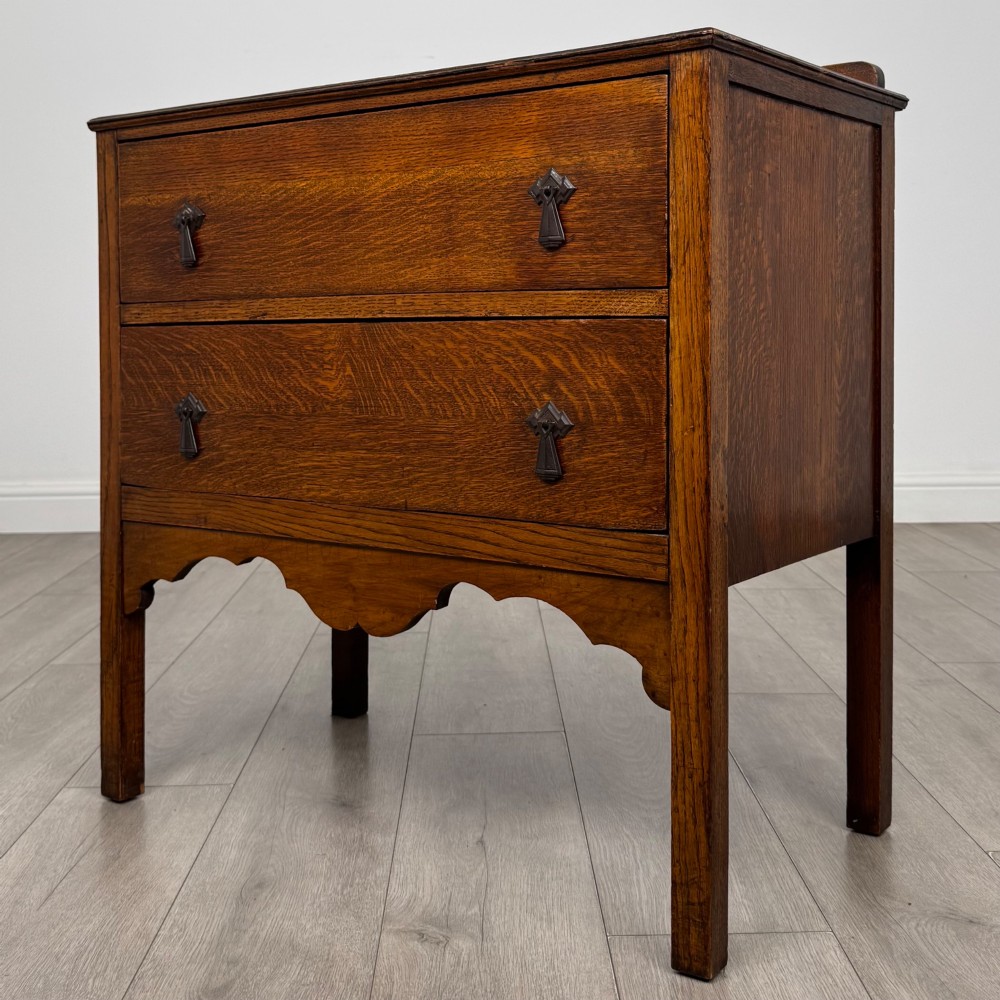 An antique golden oak chest of drawers featuring two wide drawers with stylised dark metal handles and a scalloped lower apron. View II