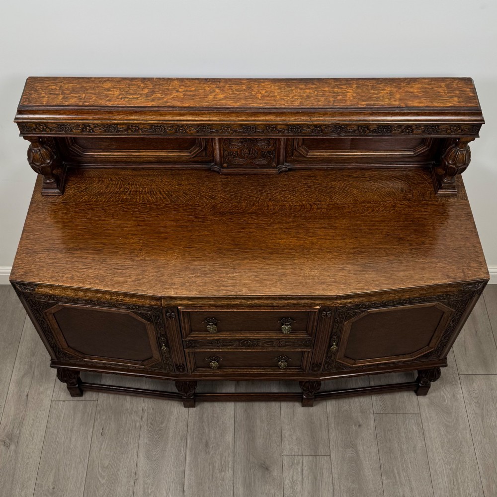 A dark carved oak sideboard featuring a raised gallery back, geometric octagonal panel doors, two central drawers with brass handles, and bulbous turned legs. View X