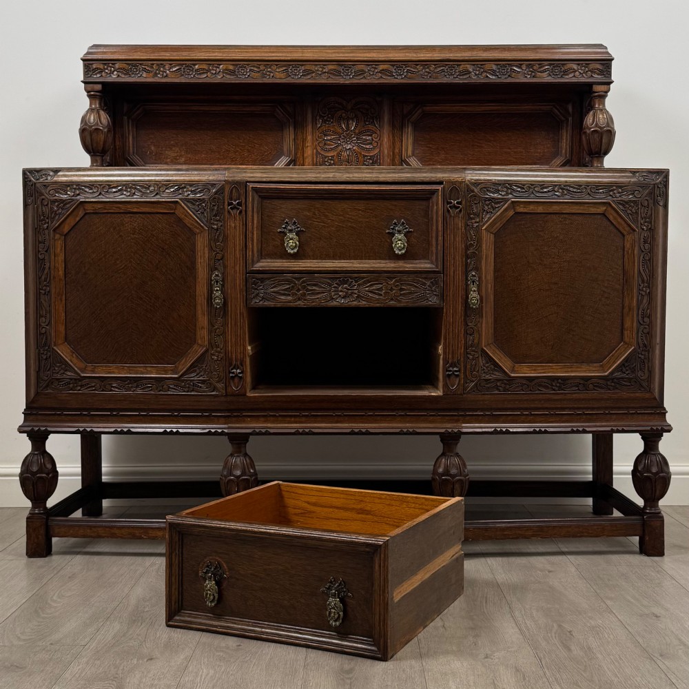 A dark carved oak sideboard featuring a raised gallery back, geometric octagonal panel doors, two central drawers with brass handles, and bulbous turned legs. View XI