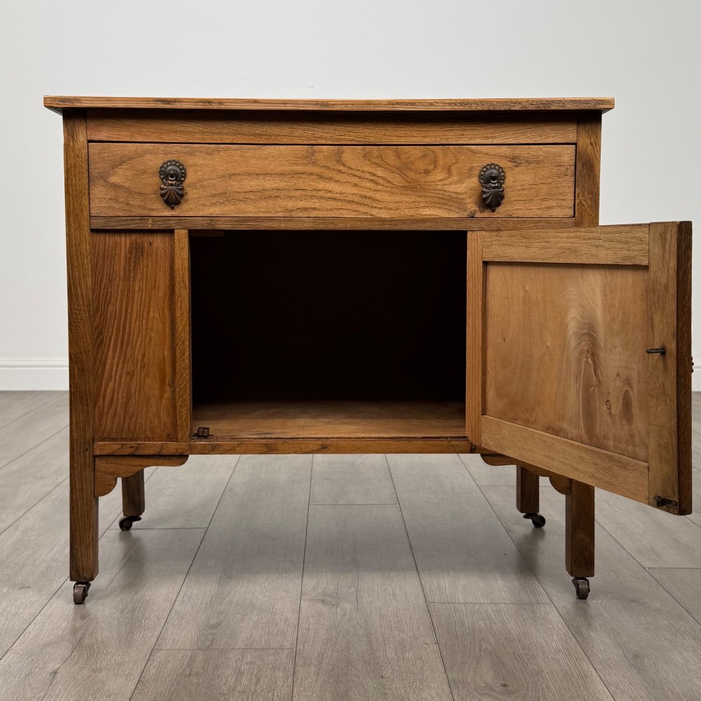 An early 20th-century golden oak side cabinet featuring a top drawer with ornate metal drop handles above a central panelled cupboard door on square legs with castors. View II