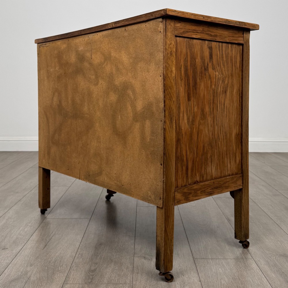 An early 20th-century golden oak side cabinet featuring a top drawer with ornate metal drop handles above a central panelled cupboard door on square legs with castors. View VII