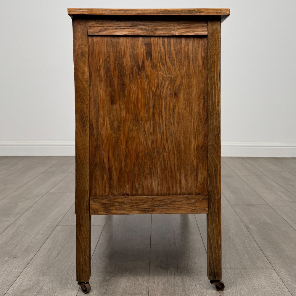 An early 20th-century golden oak side cabinet featuring a top drawer with ornate metal drop handles above a central panelled cupboard door on square legs with castors. View VIII