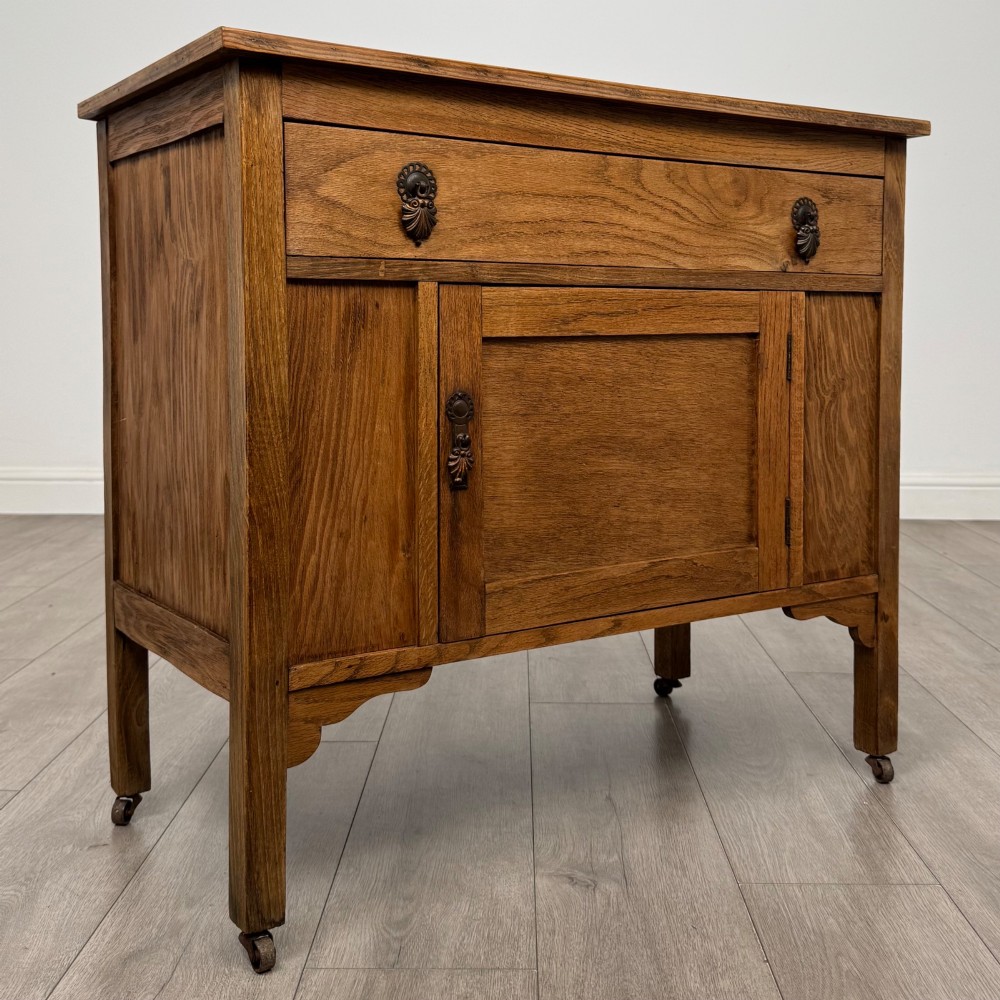 An early 20th-century golden oak side cabinet featuring a top drawer with ornate metal drop handles above a central panelled cupboard door on square legs with castors. View IX