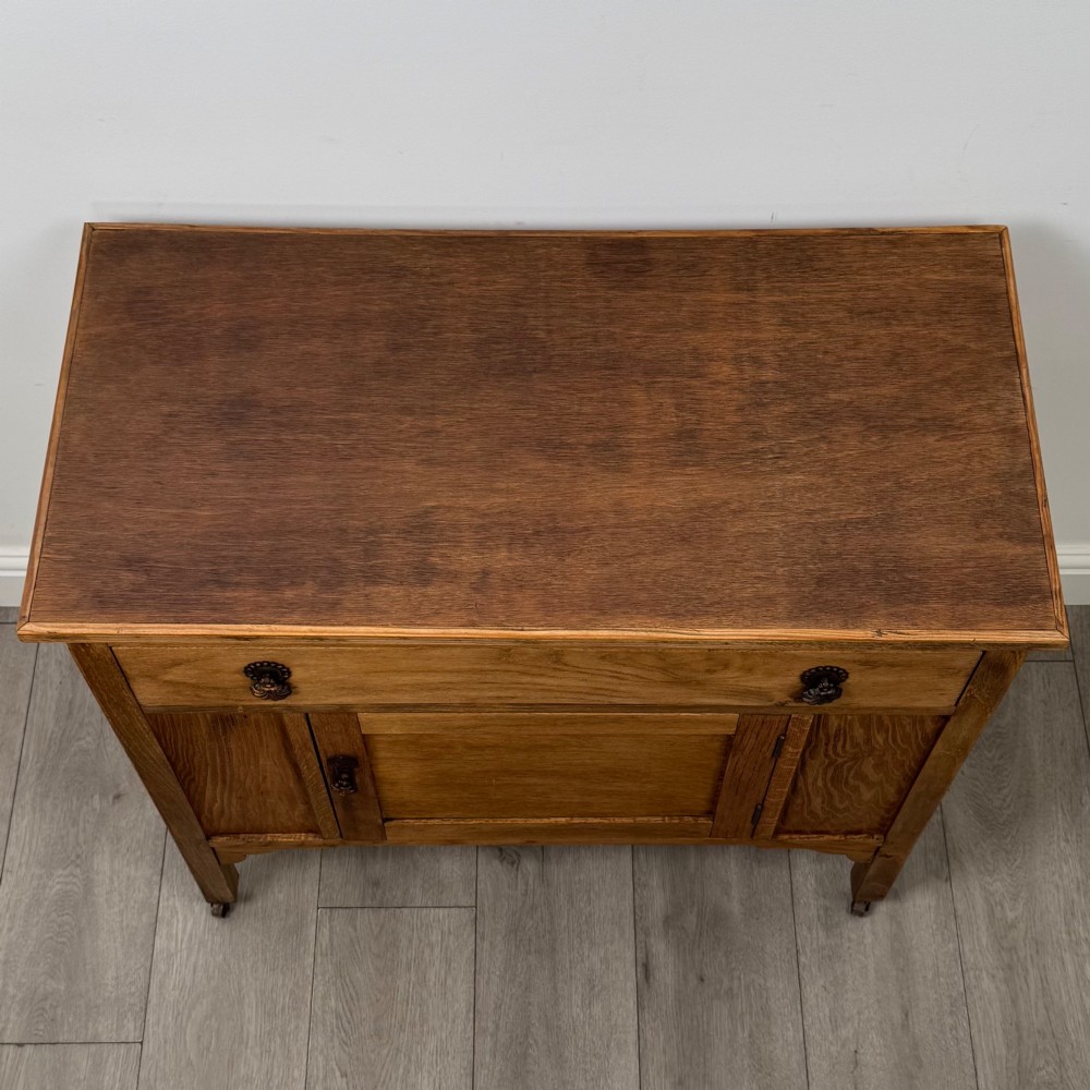 An early 20th-century golden oak side cabinet featuring a top drawer with ornate metal drop handles above a central panelled cupboard door on square legs with castors. View X