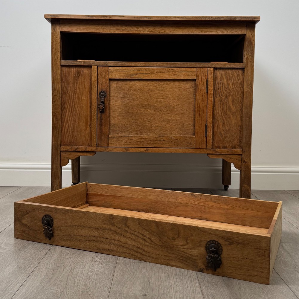 An early 20th-century golden oak side cabinet featuring a top drawer with ornate metal drop handles above a central panelled cupboard door on square legs with castors. View XI