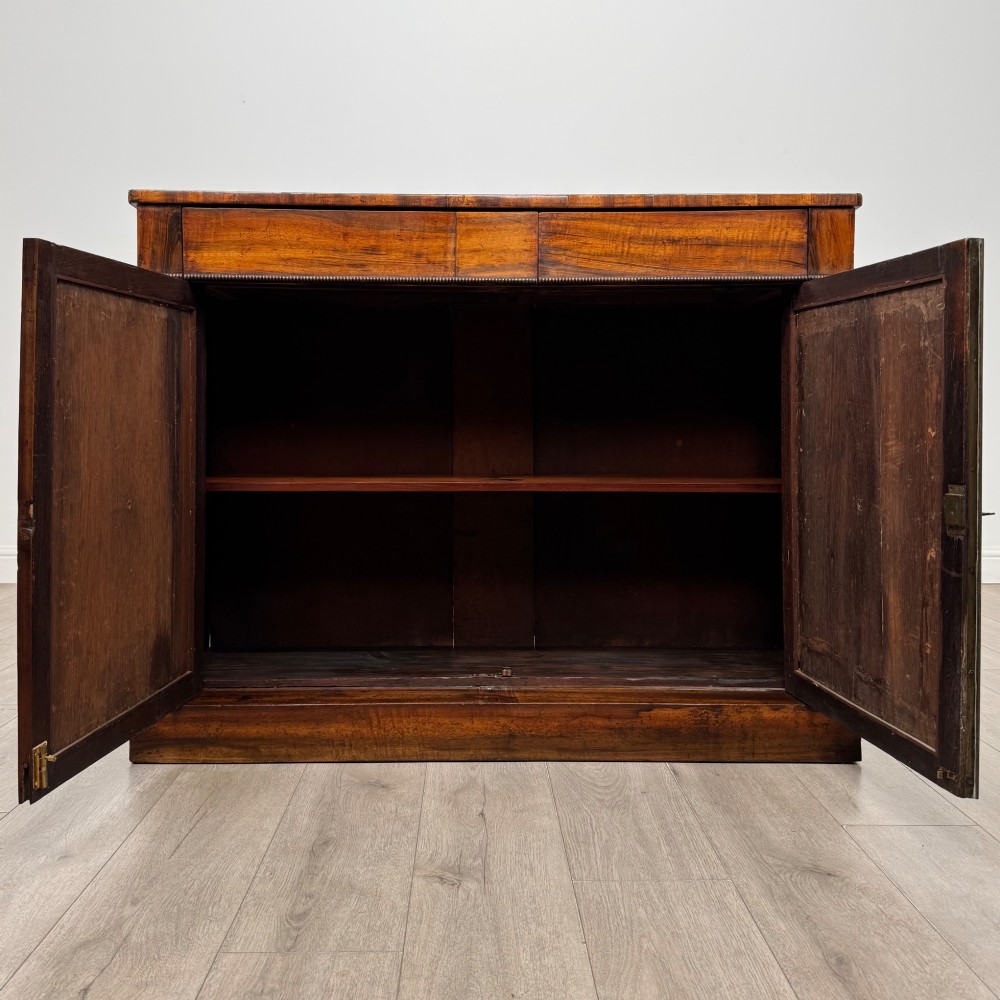 A 19th-century Regency mahogany cupboard featuring two frieze drawers, full-length turned flanking columns, and two lower doors set with pleated gold-tone fabric panels on a solid plinth base. View II