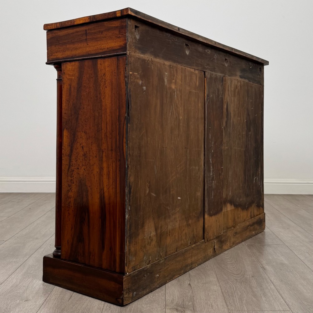 A 19th-century Regency mahogany cupboard featuring two frieze drawers, full-length turned flanking columns, and two lower doors set with pleated gold-tone fabric panels on a solid plinth base. View V
