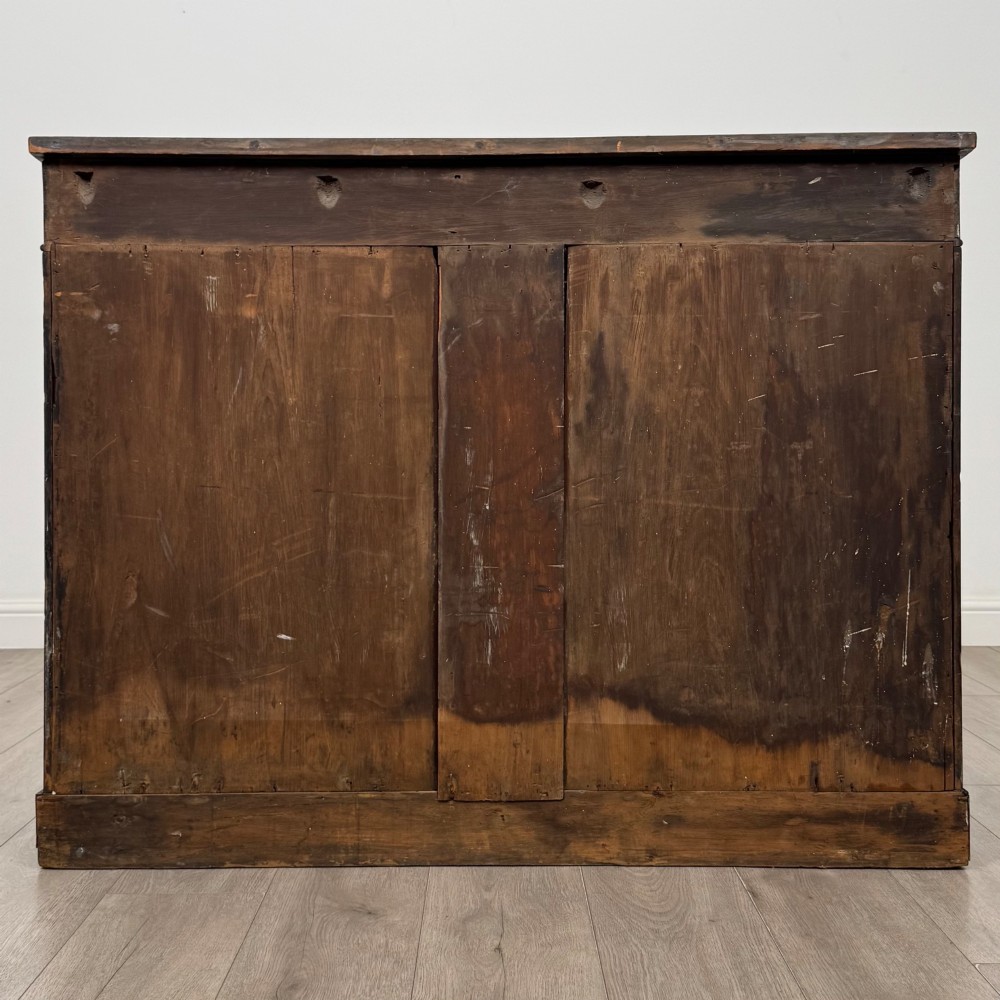 A 19th-century Regency mahogany cupboard featuring two frieze drawers, full-length turned flanking columns, and two lower doors set with pleated gold-tone fabric panels on a solid plinth base. View VI