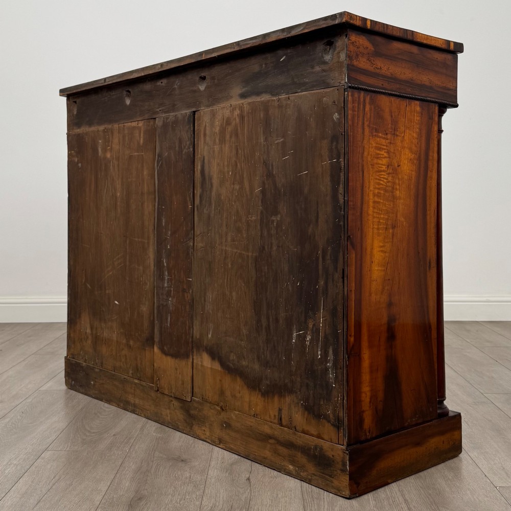 A 19th-century Regency mahogany cupboard featuring two frieze drawers, full-length turned flanking columns, and two lower doors set with pleated gold-tone fabric panels on a solid plinth base. View VII