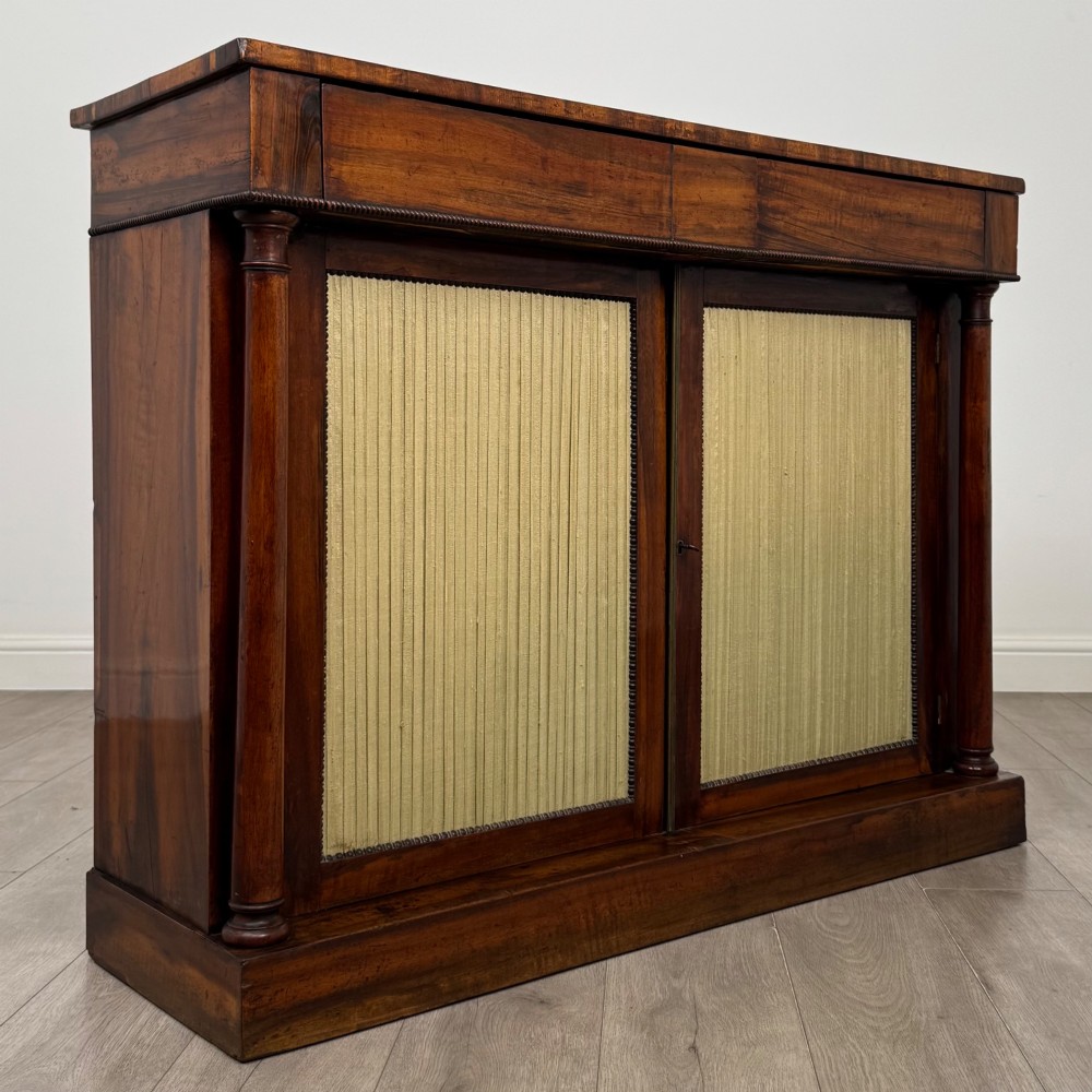 A 19th-century Regency mahogany cupboard featuring two frieze drawers, full-length turned flanking columns, and two lower doors set with pleated gold-tone fabric panels on a solid plinth base. View IX
