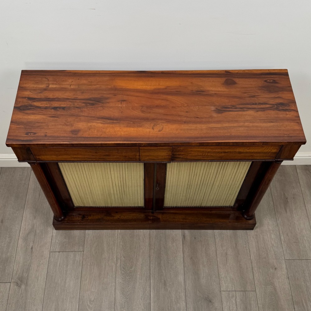 A 19th-century Regency mahogany cupboard featuring two frieze drawers, full-length turned flanking columns, and two lower doors set with pleated gold-tone fabric panels on a solid plinth base. View X