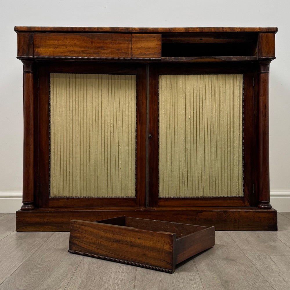 A 19th-century Regency mahogany cupboard featuring two frieze drawers, full-length turned flanking columns, and two lower doors set with pleated gold-tone fabric panels on a solid plinth base. View XI