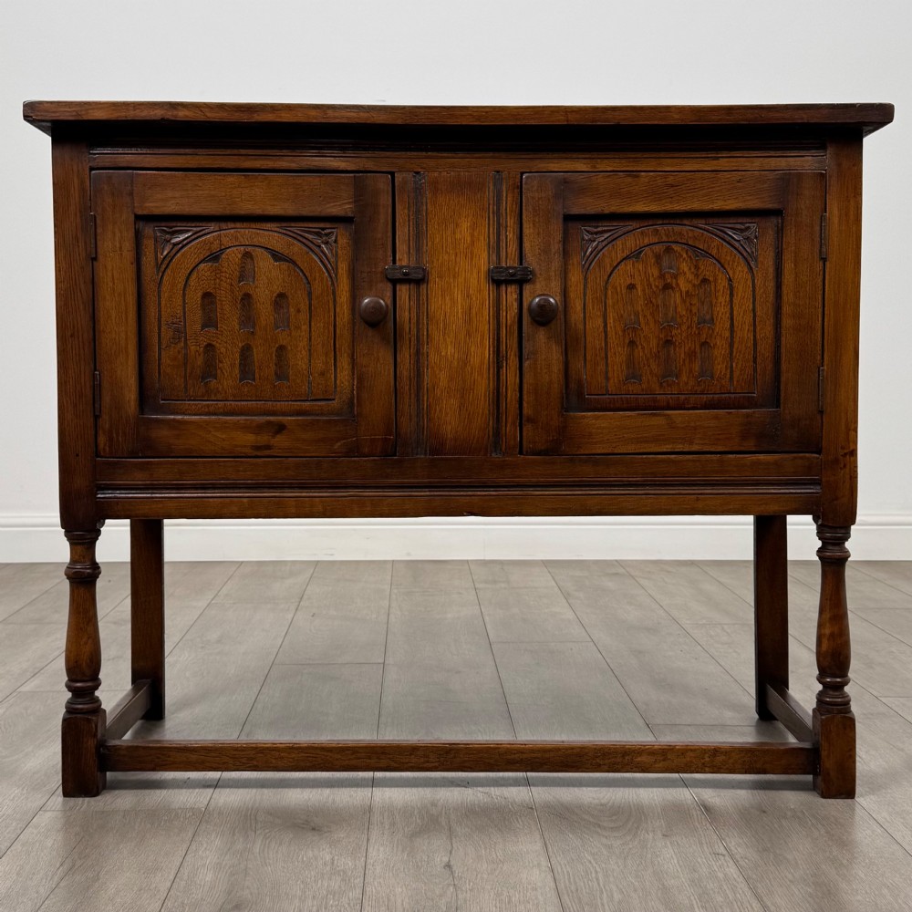 A dark oak cupboard featuring two door panels with carved Gothic arch motifs, wooden turn latches, and turned front legs connected by a perimeter stretcher.