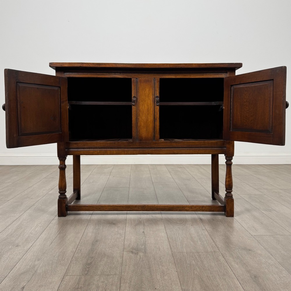 A dark oak cupboard featuring two door panels with carved Gothic arch motifs, wooden turn latches, and turned front legs connected by a perimeter stretcher. View II