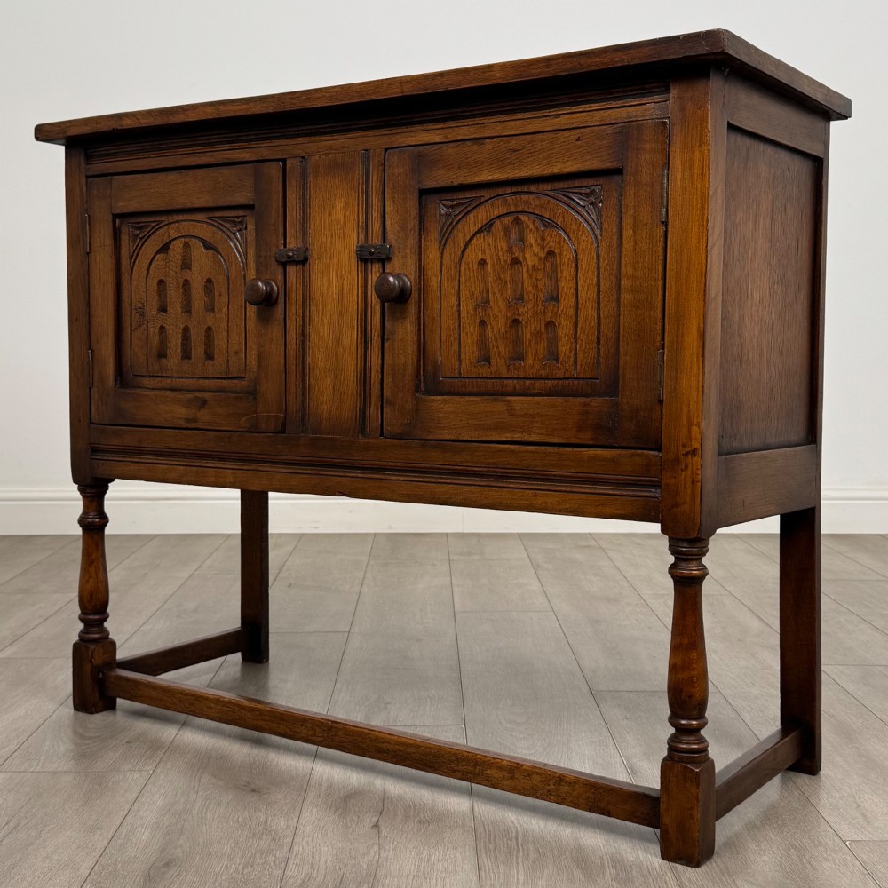 A dark oak cupboard featuring two door panels with carved Gothic arch motifs, wooden turn latches, and turned front legs connected by a perimeter stretcher. View III