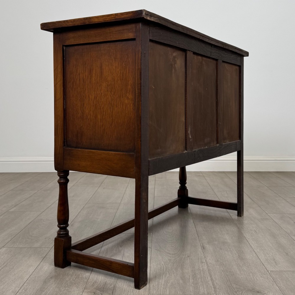 A dark oak cupboard featuring two door panels with carved Gothic arch motifs, wooden turn latches, and turned front legs connected by a perimeter stretcher. View V