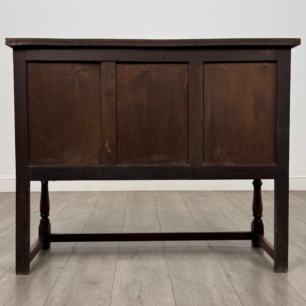 A dark oak cupboard featuring two door panels with carved Gothic arch motifs, wooden turn latches, and turned front legs connected by a perimeter stretcher. View VI