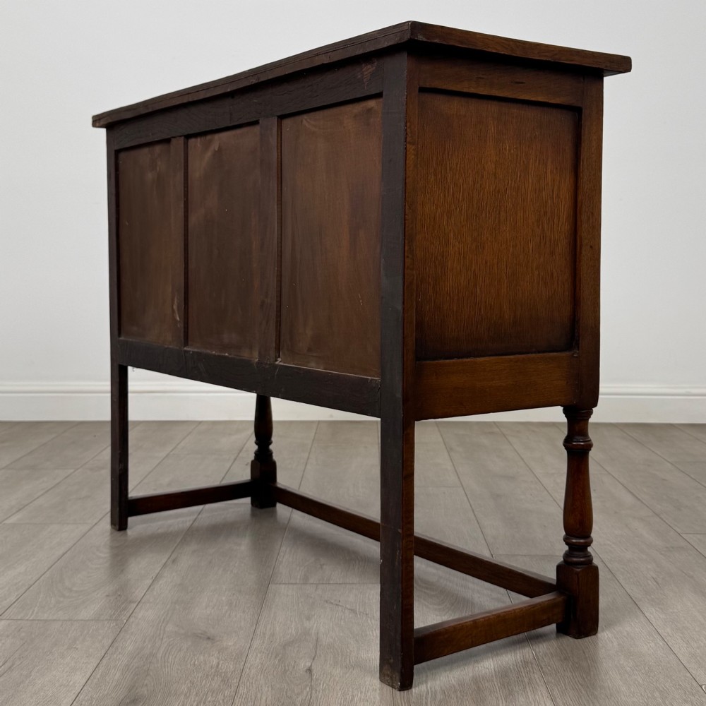 A dark oak cupboard featuring two door panels with carved Gothic arch motifs, wooden turn latches, and turned front legs connected by a perimeter stretcher. View VII