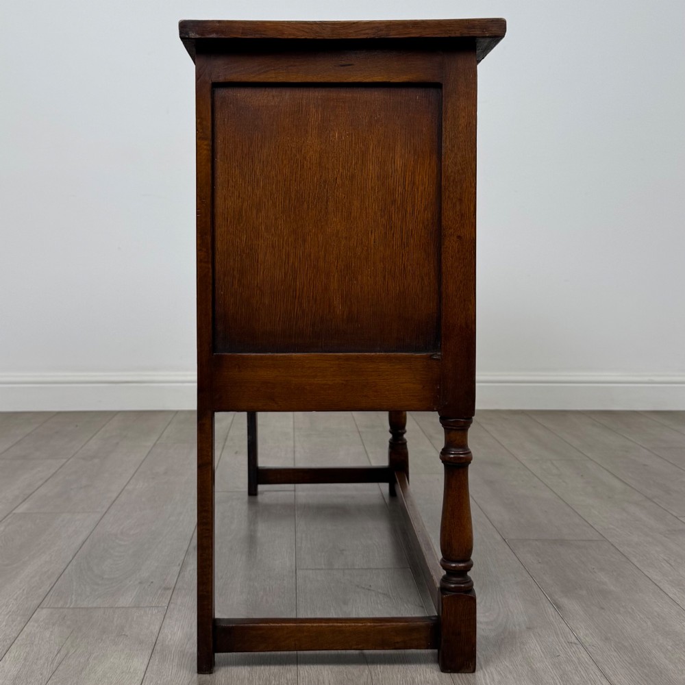A dark oak cupboard featuring two door panels with carved Gothic arch motifs, wooden turn latches, and turned front legs connected by a perimeter stretcher. View VIII