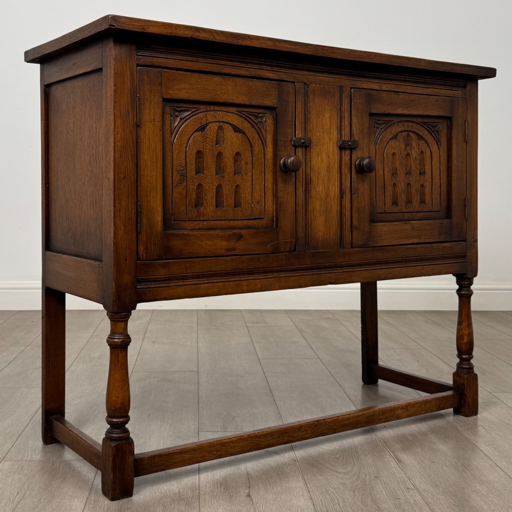 A dark oak cupboard featuring two door panels with carved Gothic arch motifs, wooden turn latches, and turned front legs connected by a perimeter stretcher. View IX
