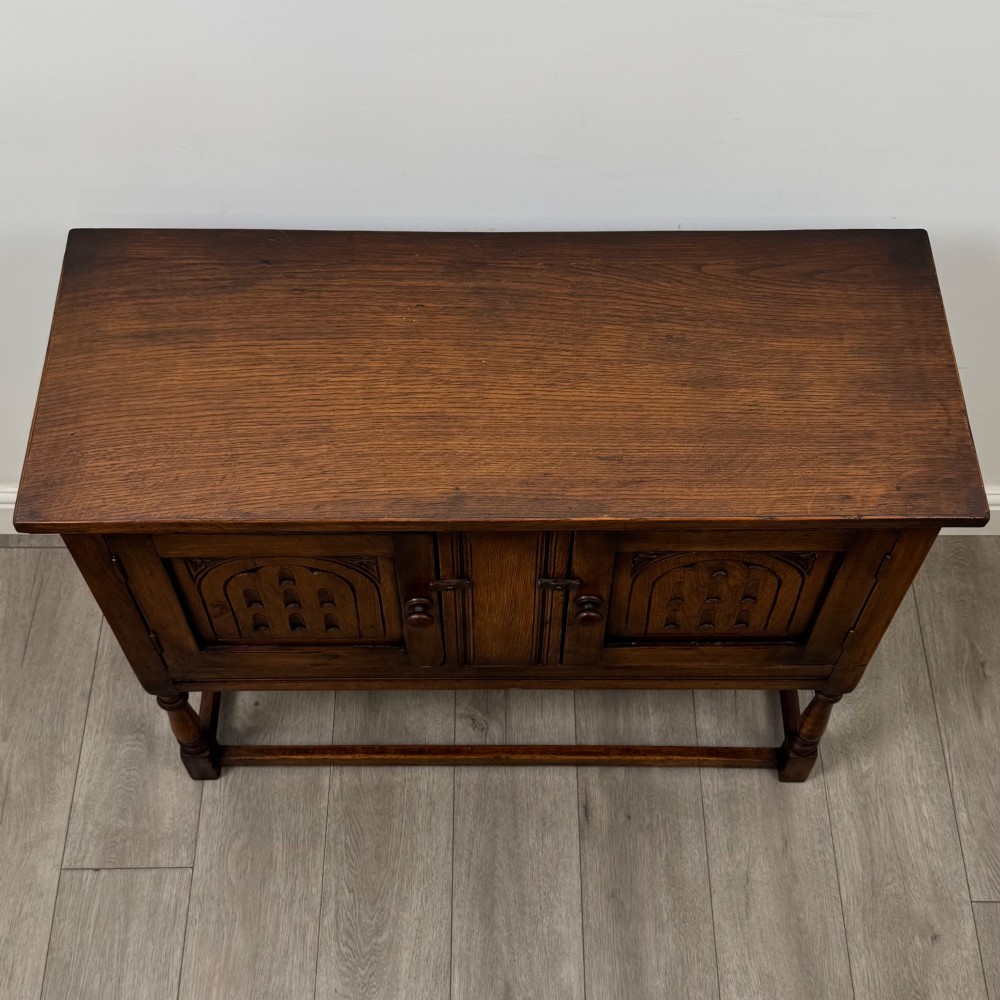 A dark oak cupboard featuring two door panels with carved Gothic arch motifs, wooden turn latches, and turned front legs connected by a perimeter stretcher. View X