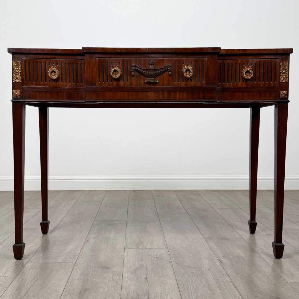 A dark mahogany 20th-century Adam style side table featuring a central carved urn motif, fluted frieze drawers, brass ring handles, and square tapered legs ending in spade feet.