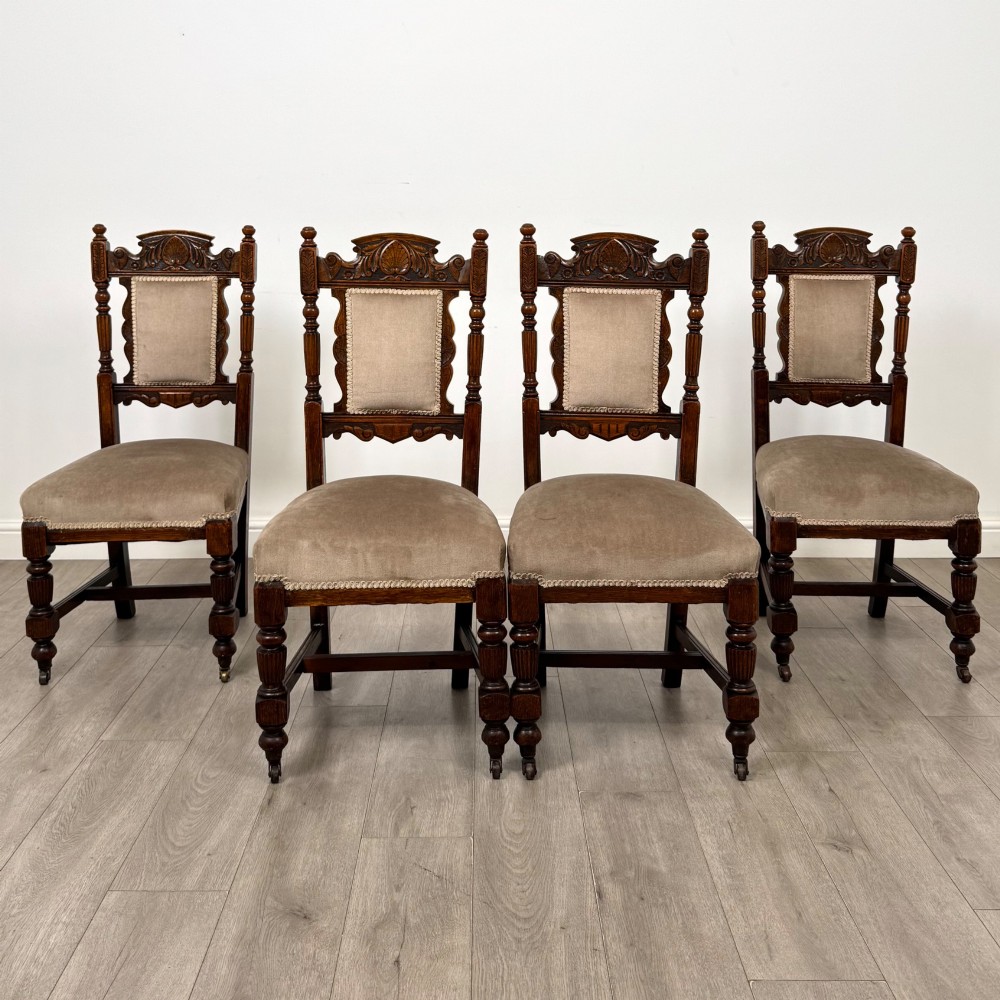 A set of four 19th-century Victorian carved oak dining chairs featuring beige upholstered seats, turned legs, and detailed crest carved backrests.