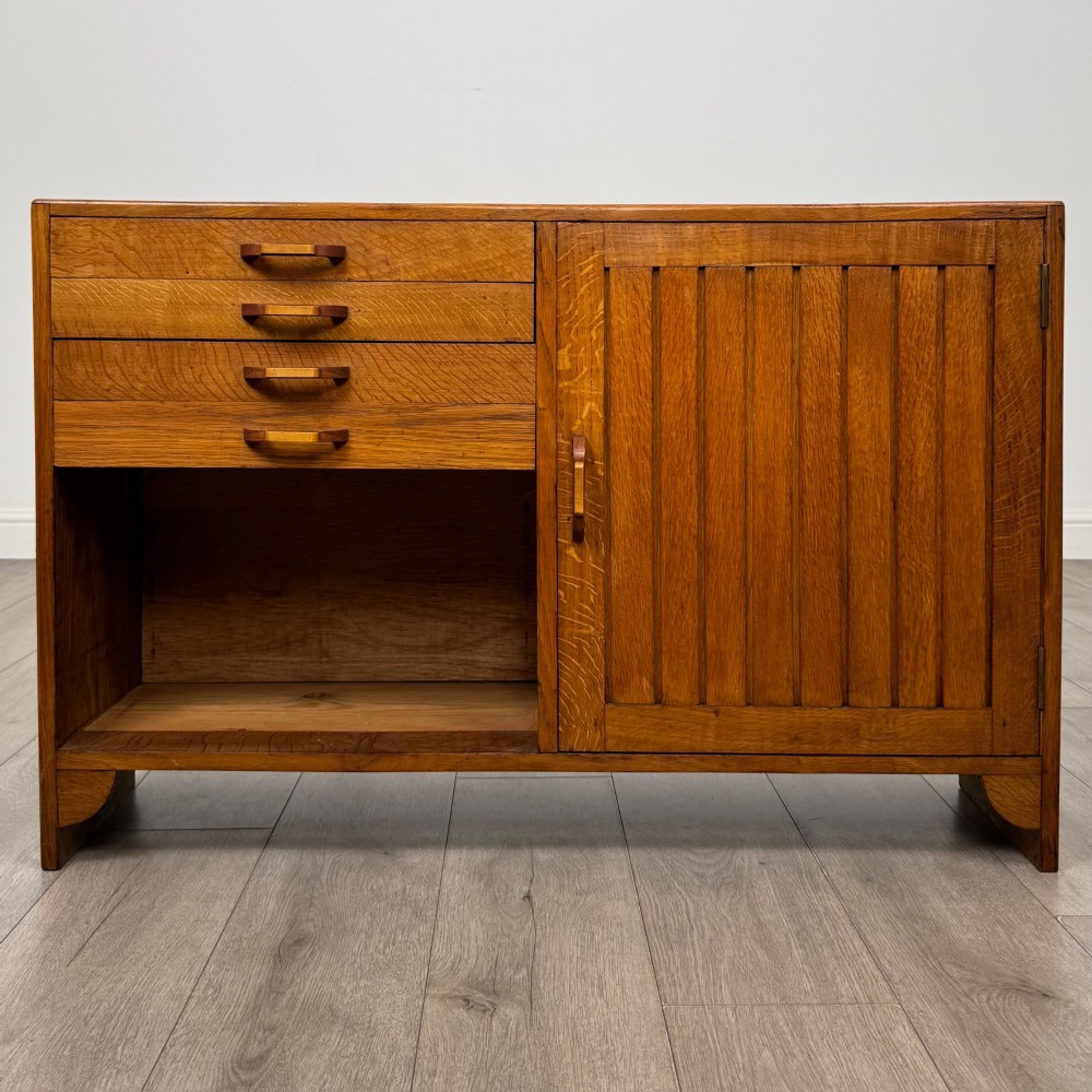 A compact golden oak sideboard featuring four shallow drawers on the left, an open shelf below, and a slatted single door cabinet on the right.