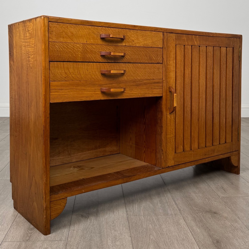 A compact golden oak sideboard featuring four shallow drawers on the left, an open shelf below, and a slatted single door cabinet on the right. View IX