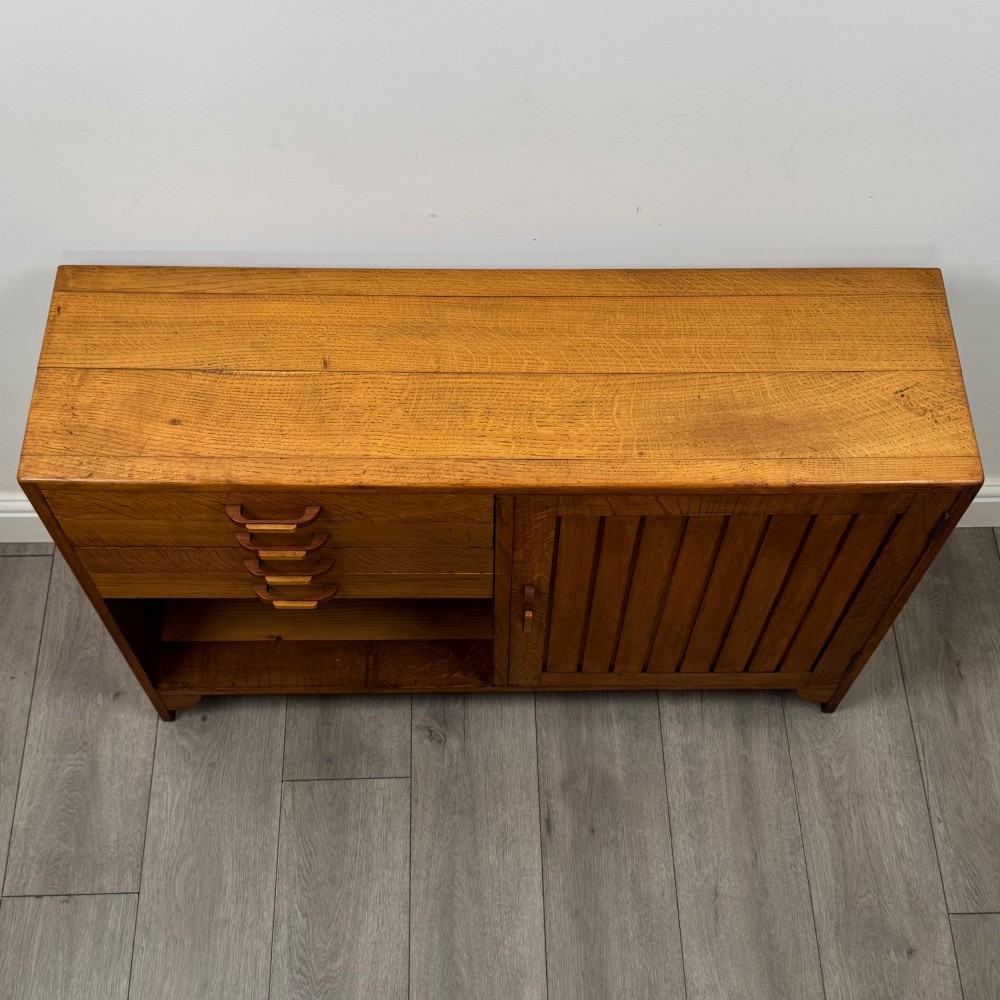 A compact golden oak sideboard featuring four shallow drawers on the left, an open shelf below, and a slatted single door cabinet on the right. View X