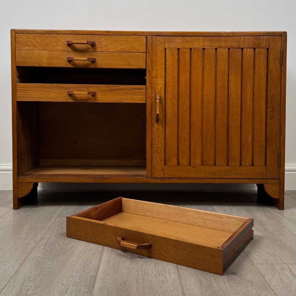A compact golden oak sideboard featuring four shallow drawers on the left, an open shelf below, and a slatted single door cabinet on the right. View XI