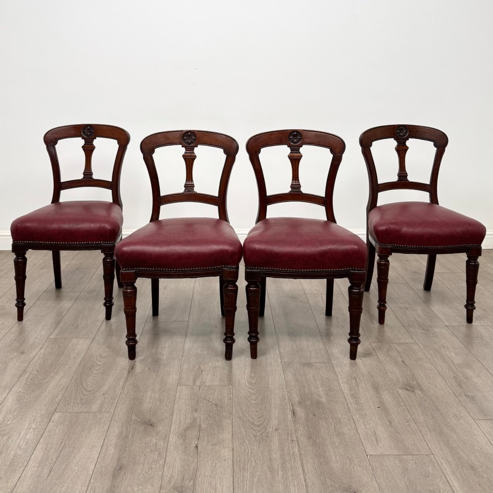 A set of four 19th-century mahogany dining chairs featuring carved open backs, dark red leather upholstered seats with stud trim, and turned front legs.
