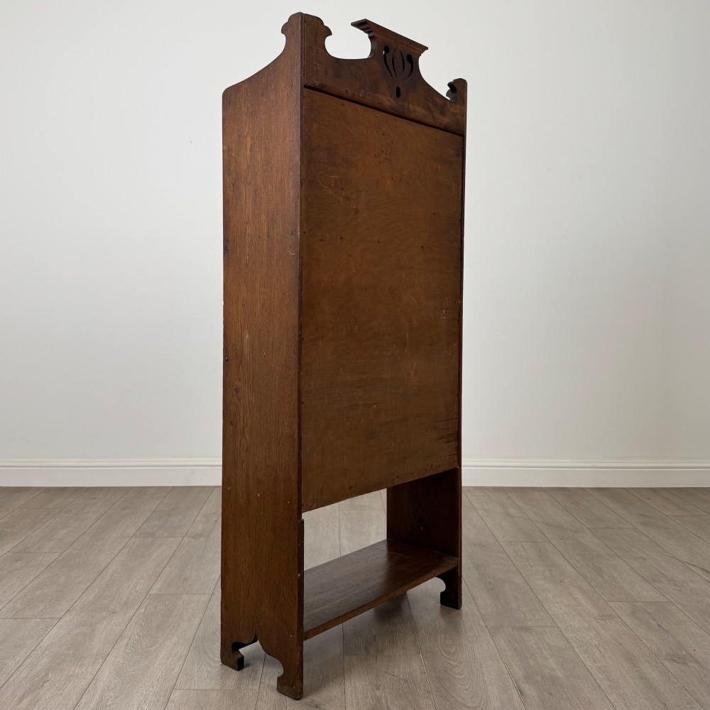 An Arts and Crafts dark oak writing bureau featuring a top crest with a pierced heart motif, a small upper cupboard with a leaded oval window, a fall-front writing section, and two open lower shelves. View V