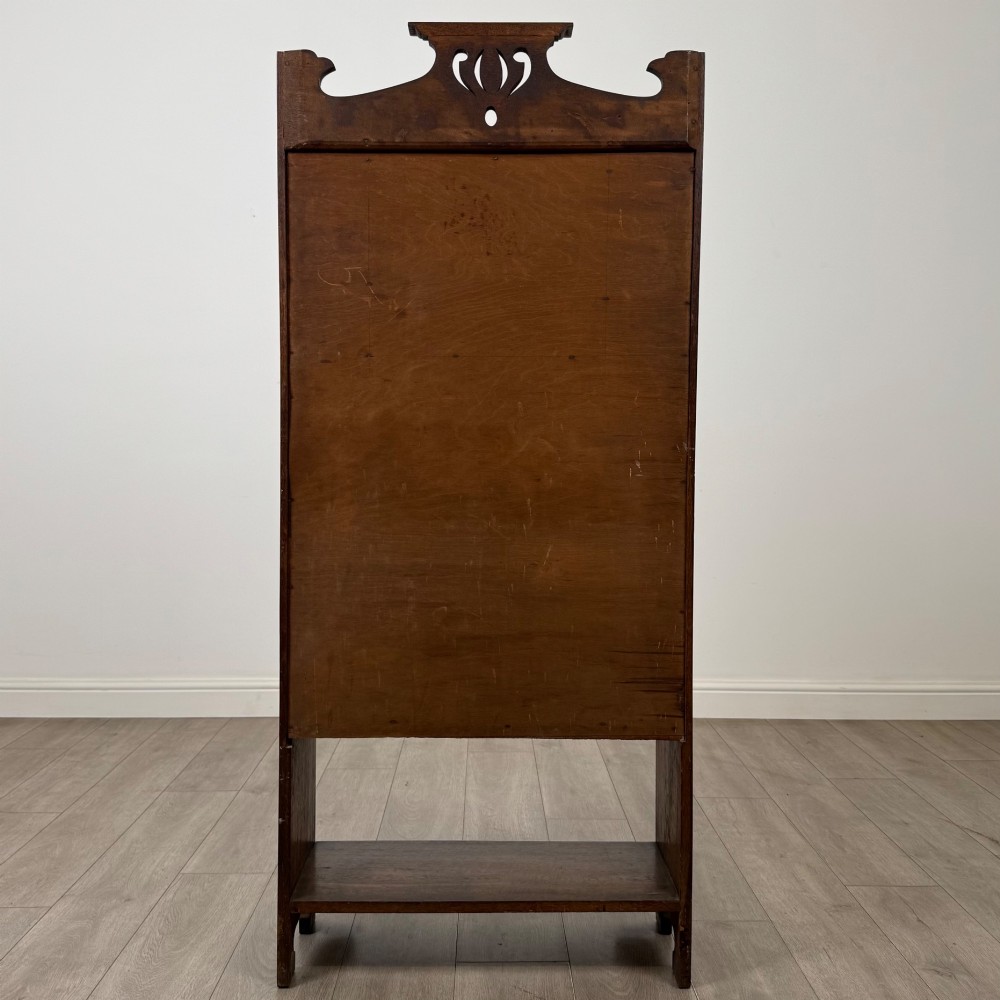 An Arts and Crafts dark oak writing bureau featuring a top crest with a pierced heart motif, a small upper cupboard with a leaded oval window, a fall-front writing section, and two open lower shelves. View VI