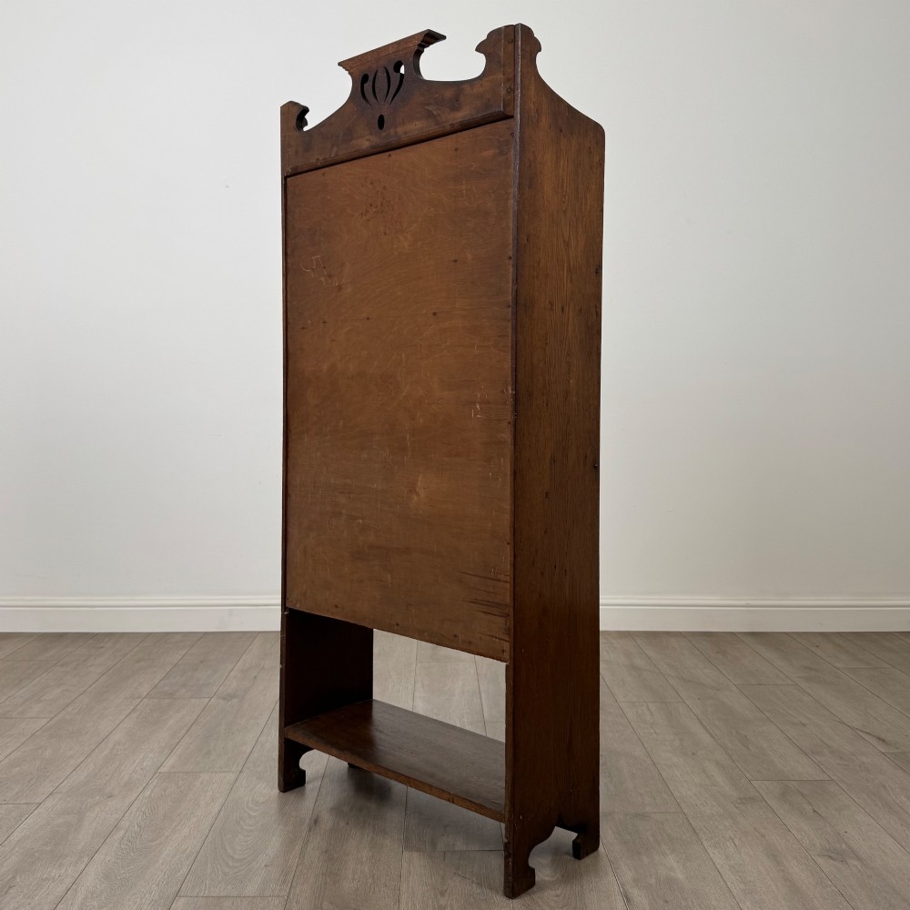 An Arts and Crafts dark oak writing bureau featuring a top crest with a pierced heart motif, a small upper cupboard with a leaded oval window, a fall-front writing section, and two open lower shelves. View VII