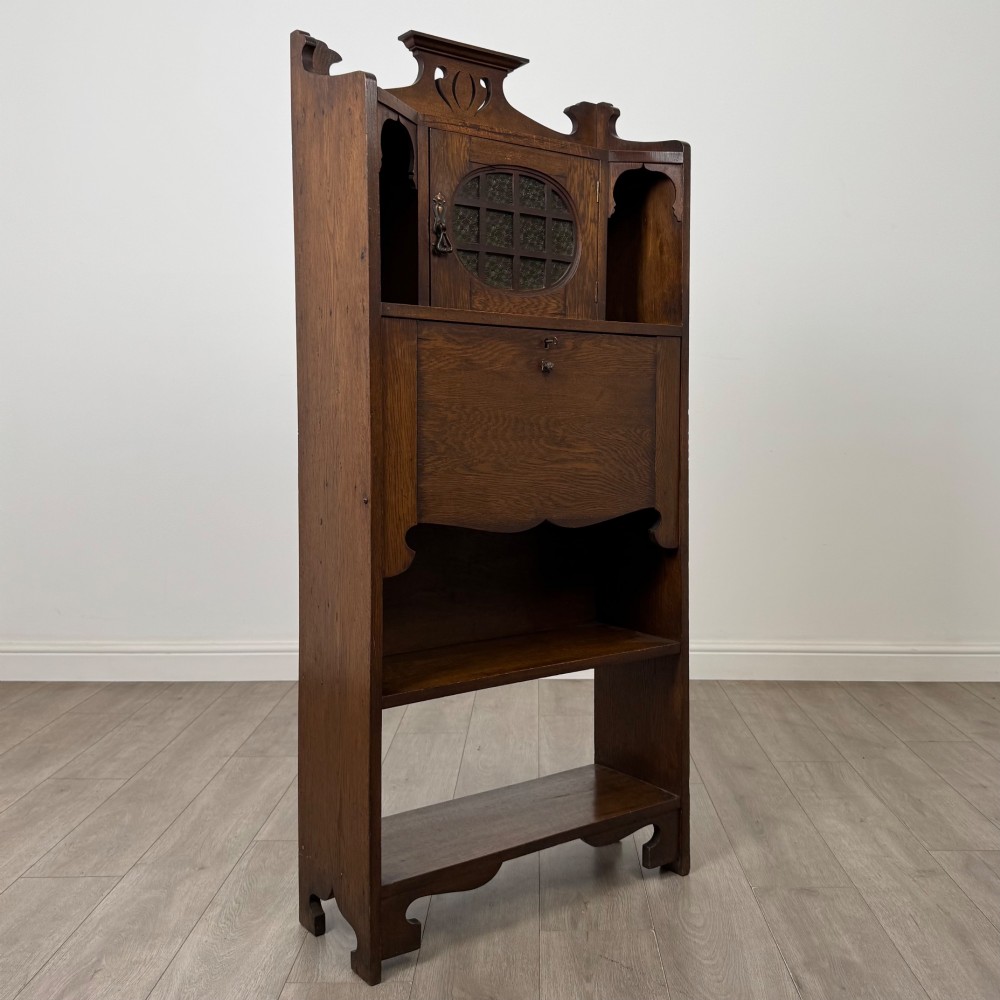 An Arts and Crafts dark oak writing bureau featuring a top crest with a pierced heart motif, a small upper cupboard with a leaded oval window, a fall-front writing section, and two open lower shelves. View IX