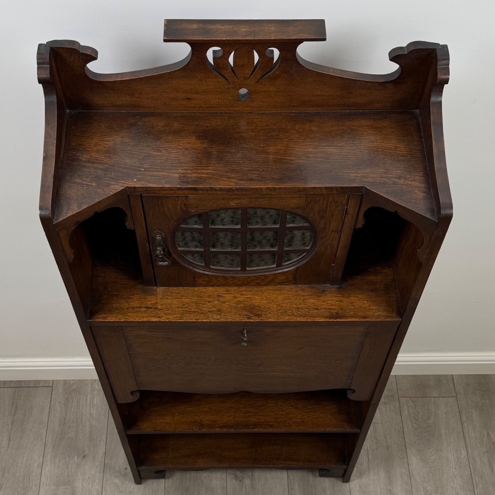 An Arts and Crafts dark oak writing bureau featuring a top crest with a pierced heart motif, a small upper cupboard with a leaded oval window, a fall-front writing section, and two open lower shelves. View X