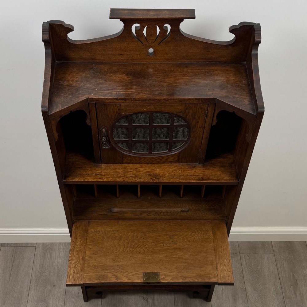 An Arts and Crafts dark oak writing bureau featuring a top crest with a pierced heart motif, a small upper cupboard with a leaded oval window, a fall-front writing section, and two open lower shelves. View XI