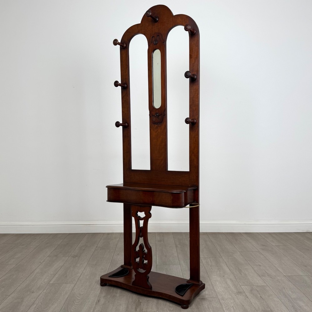 A freestanding Victorian mahogany hall stand featuring turned coat pegs, a narrow central mirror panel, a small shelf, and an umbrella holder base. View II