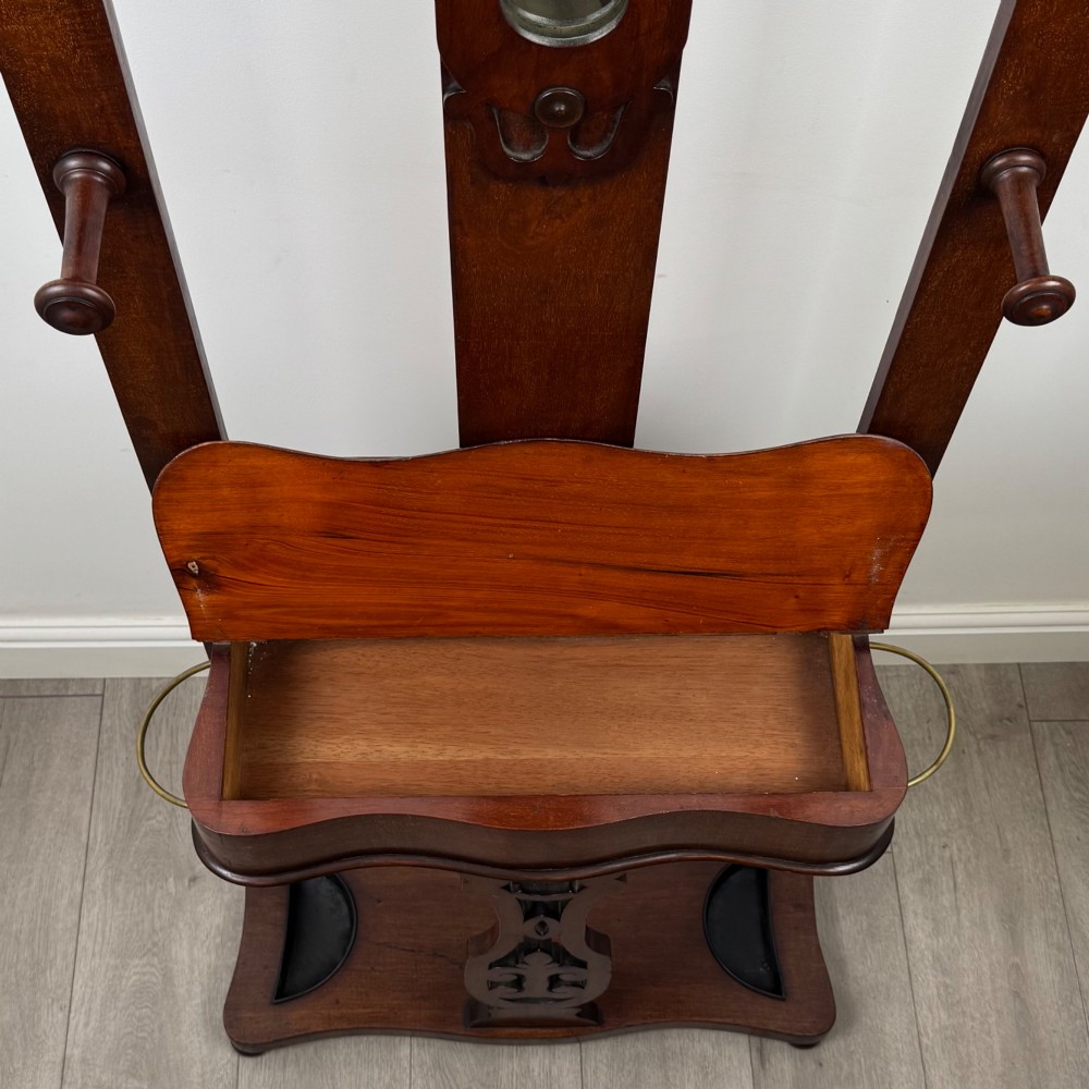 A freestanding Victorian mahogany hall stand featuring turned coat pegs, a narrow central mirror panel, a small shelf, and an umbrella holder base. View X