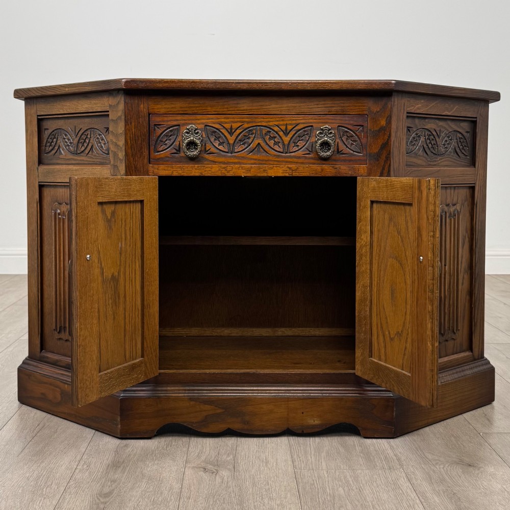 A dark oak low cupboard featuring canted corners, carved linenfold door panels, an upper drawer with carved foliage, and brass hinges. View II